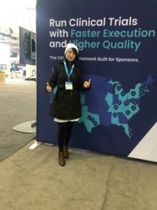 A woman participating in clinical trials with faster execution and quality stands in front of a sign that reads "AAD 2022.