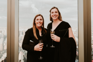 Two women holding champagne glasses in front of a city skyline at ASRS 2022.