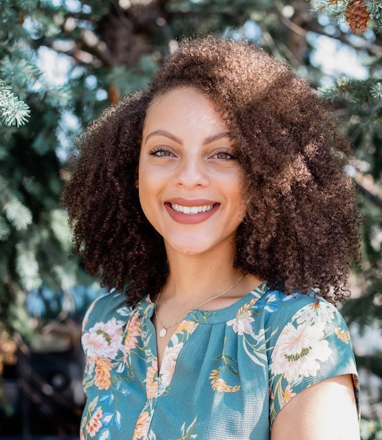 A woman with afro hair smiling in front of a tree while holding a vial.