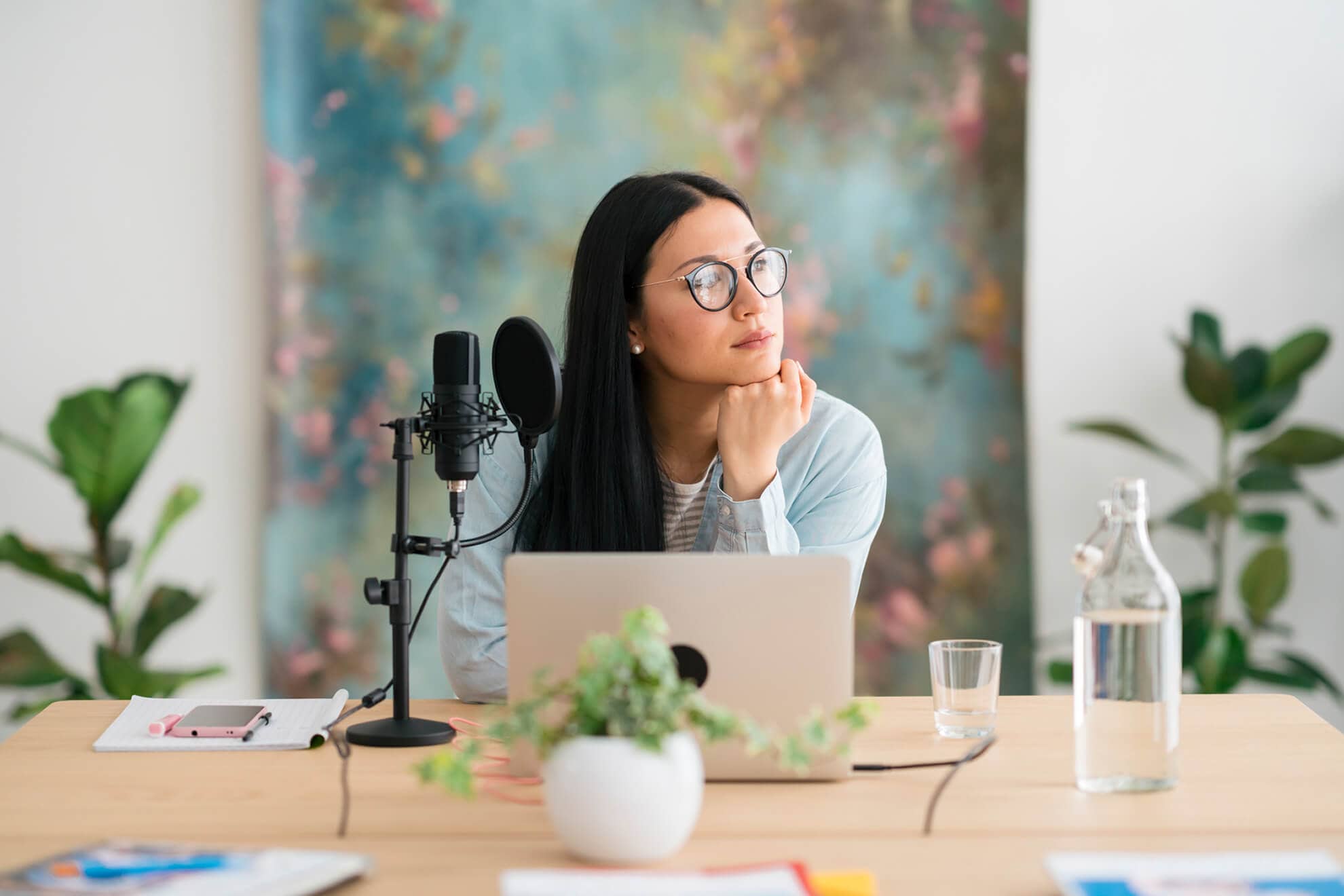 Contemplating Asian female radio host leaning on hand and looking away while sitting at table with netbook and mic prepared for recording podcast